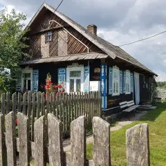Drewniany dom z niebieskimi, otwartymi okiennicami, ozdobiony ludowymi wzorami. Wokół zielony trawnik i drewniany płot.