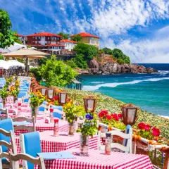 Urokliwa restauracja nad morzem w Bułgarii. Stoliki z czerwono-białymi obrusami i niebieskimi krzesłami czekają na gości, a w tle widać turkusowe morze i malownicze klify.
