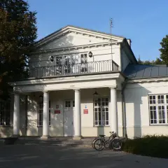Pałac Potulickich w Pruszkowie, z kolumnowym portykiem i balkonem, otoczony zielenią. Dwa rowery stoją przed wejściem.