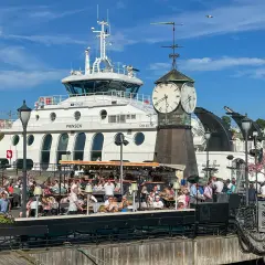 Tętniący życiem deptak w Oslo, z ludźmi siedzącymi w kawiarniach, na tle białego statku i zabytkowej wieży zegarowej. Oslo czy warto? Zdecydowanie tak!