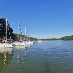Żaglówki cumują w porcie w Rucianem Nidzie. Słoneczny dzień nad jeziorem, zalesiony brzeg w oddali.