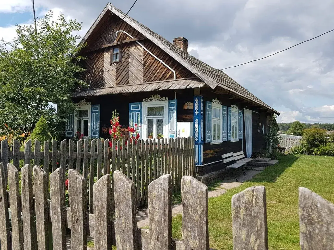 Drewniany dom z niebieskimi, otwartymi okiennicami, ozdobiony ludowymi wzorami. Wokół zielony trawnik i drewniany płot.