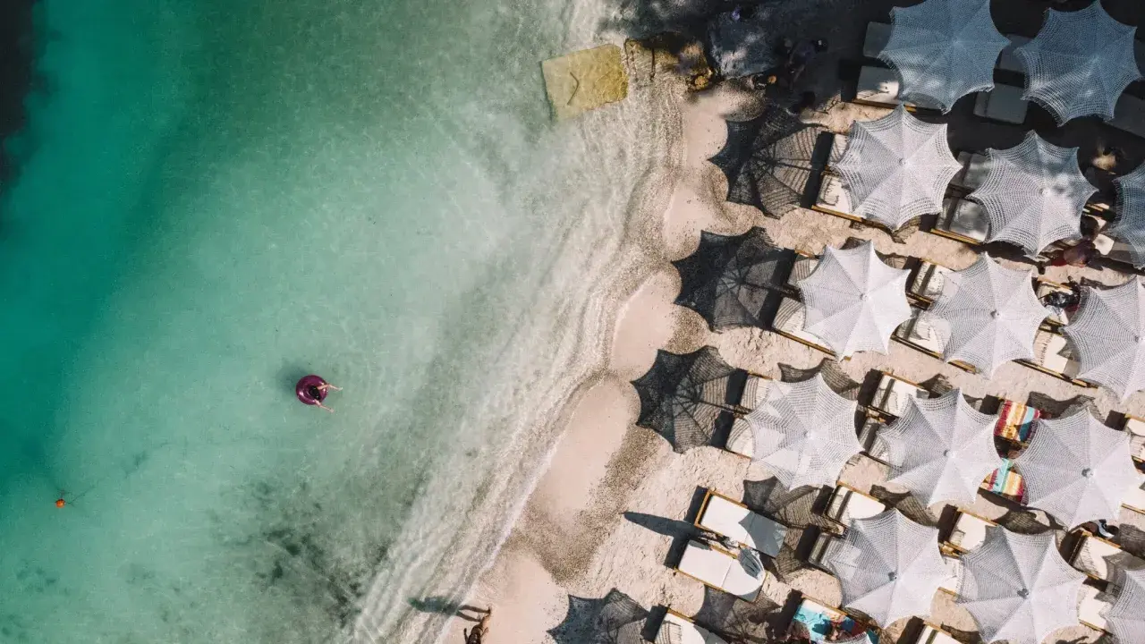 Widok z lotu ptaka na turkusowe morze i piaszczyste plaże Albanii. Jedna osoba relaksuje się na dmuchanym kole, a obok rzędy białych parasoli i leżaków.