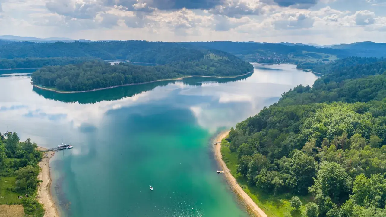 Malownicze jezioro otoczone lasami, idealne miejsce, gdzie warto pojechać nad jezioro w Polsce. Widać łodzie i piaszczystą plażę.