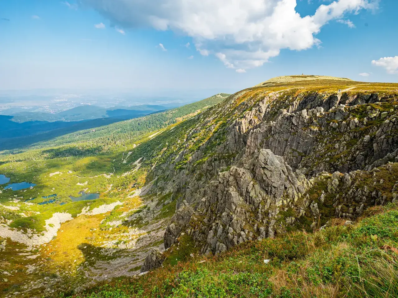Widok na skaliste zbocze i zieloną dolinę z jeziorami. W oddali widać miasto. To krajobraz, który mógłby być nazwany "śnieżne kotły".