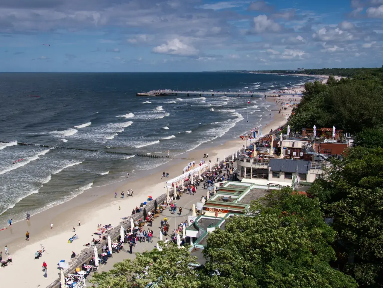 Tętniąca życiem promenada nad morzem, z plażą pełną ludzi, kawiarniami i molo w oddali.
