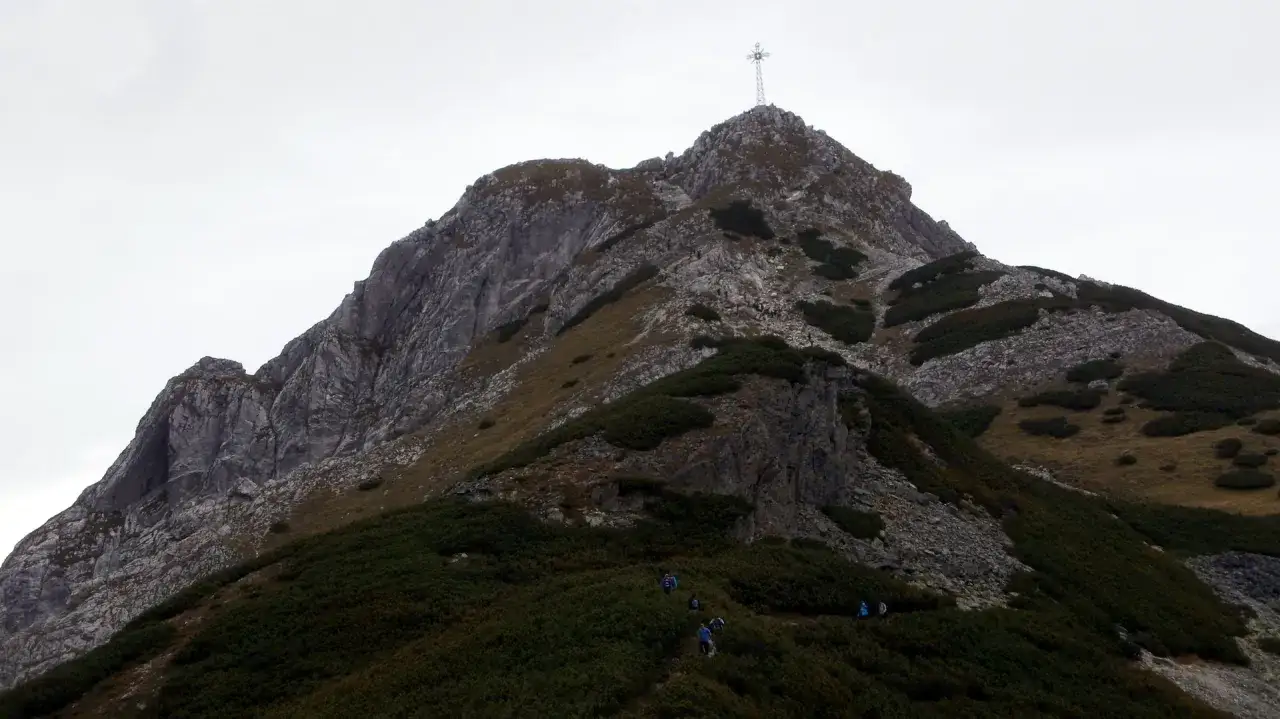 Grupa turystów podąża stromą trasą na Giewont. Widoczny szczyt z krzyżem i skaliste zbocza.