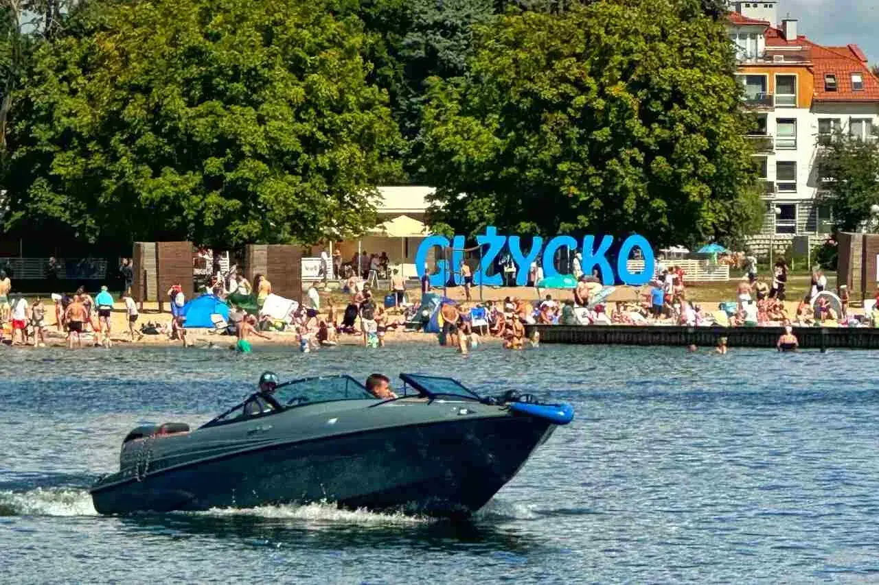 Motorówka płynie po jeziorze w Giżycku na Mazurach, gdzie na plaży wypoczywają tłumy ludzi.