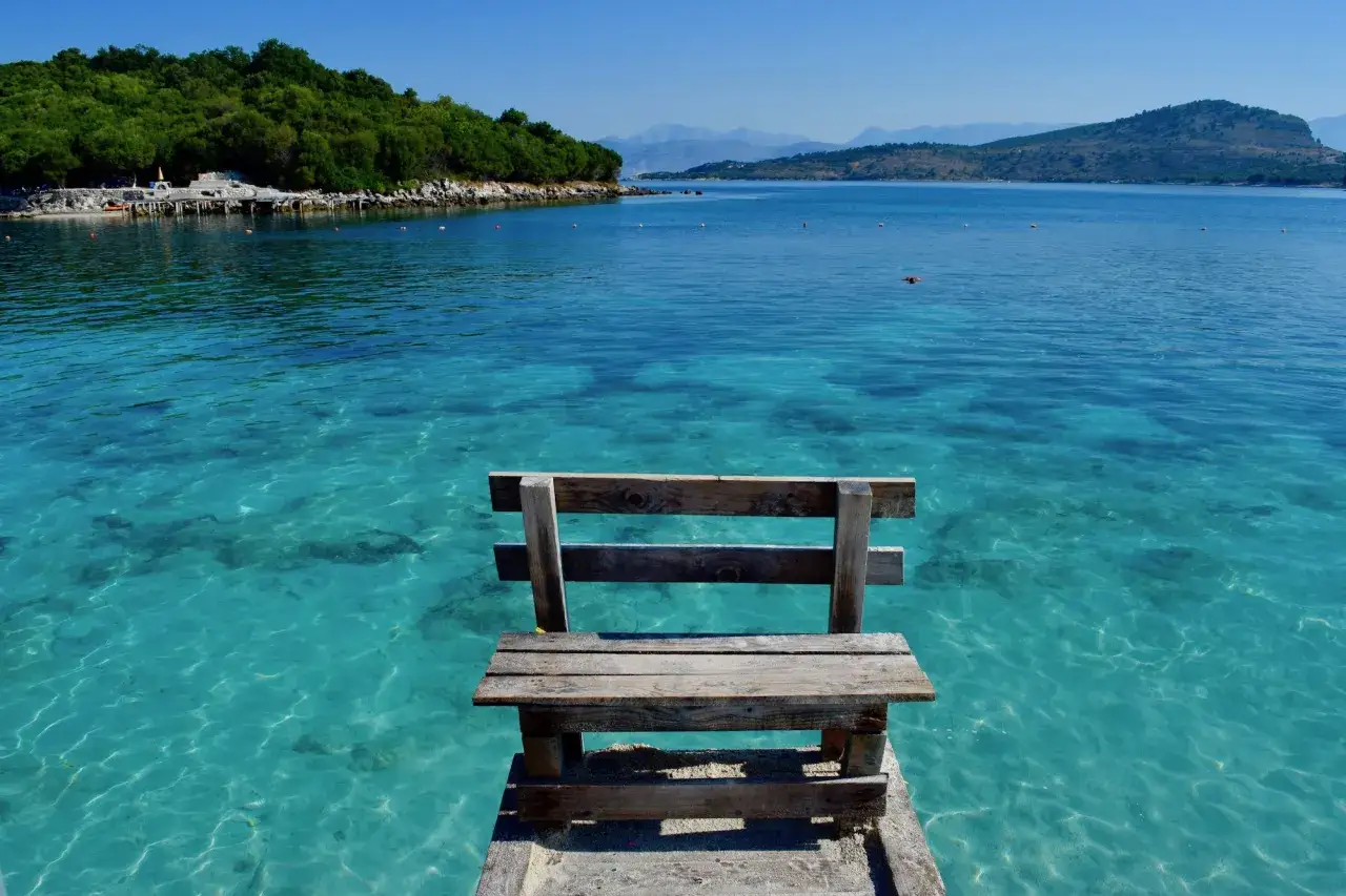 Drewniana ławka na pomoście nad krystalicznie czystą wodą. W tle zielone wzgórza i góry. Idealne miejsce na relaks na albania plaże.