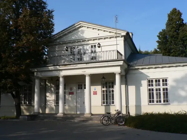 Pałac Potulickich w Pruszkowie, z kolumnowym portykiem i balkonem, otoczony zielenią. Dwa rowery stoją przed wejściem.
